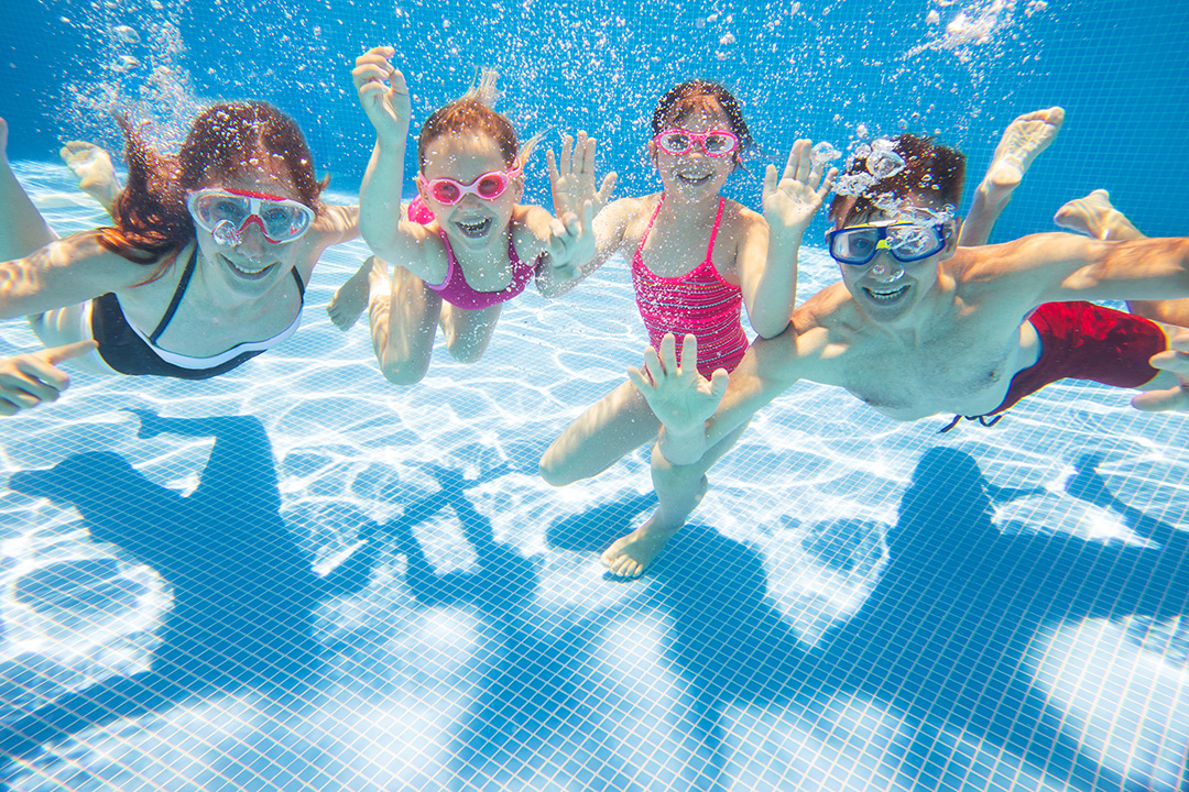 four children of various ages underwater in a pool, waving at the camera